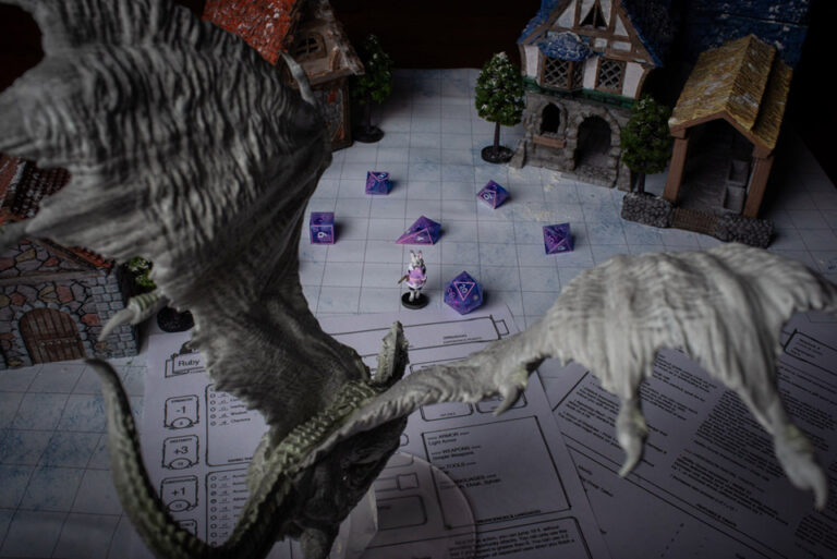 A set of purple and pink D&D dice with a matching Harengon bunny miniature character, in front of a model scene of a large dragon in a village.