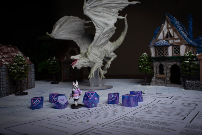 A set of purple and pink D&D dice with a matching Harengon bunny miniature character, in front of a model scene of a large dragon in a village.