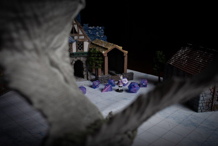A set of purple and pink D&D dice with a matching Harengon bunny miniature character, in front of a model scene of a large dragon in a village.